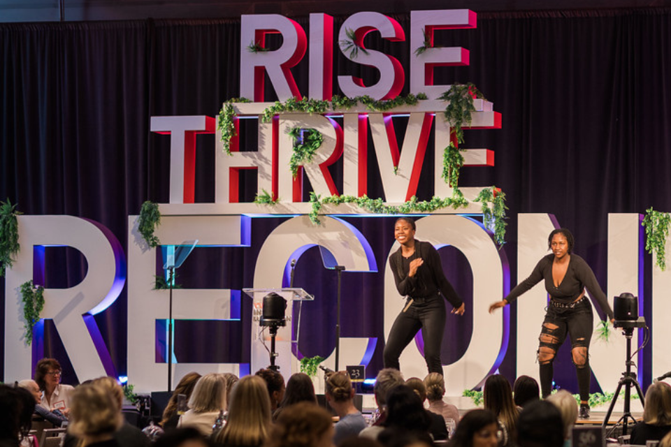 Two young black women standing in front of a logo on stage saying "Rise thrive reconnect"