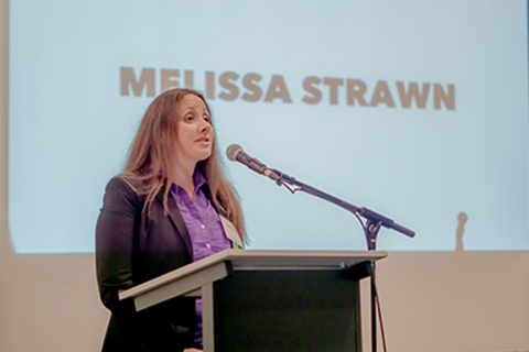 Picture of client speaker on stage with "Melissa Strawn" on projector screen behind her