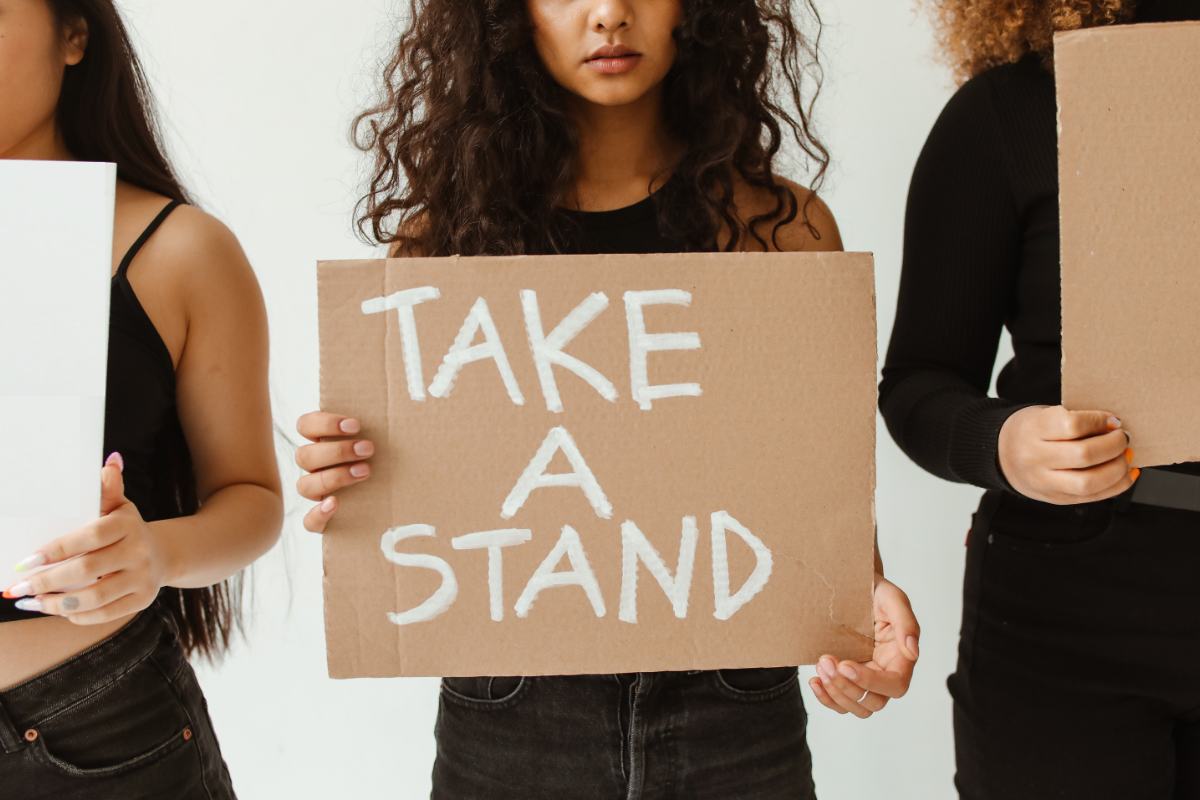 Three women holding signs, the middle one reading "take a stand"