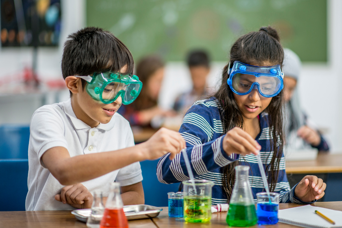 A photo of a young boy and a young girl wearing goggles while using pipettes to transfer colored water into glass beakers.