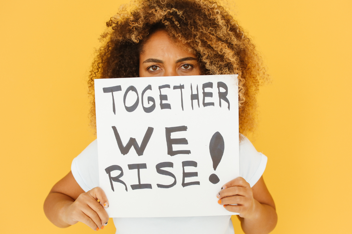 Woman standing against a yellow background holding a sign that says "Together we rise!"