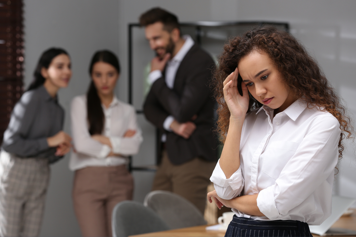 Photo of four people in an office space. Three are standing in the back looking at the person in the forefront, who has their hand to their temple and looks frustrated.