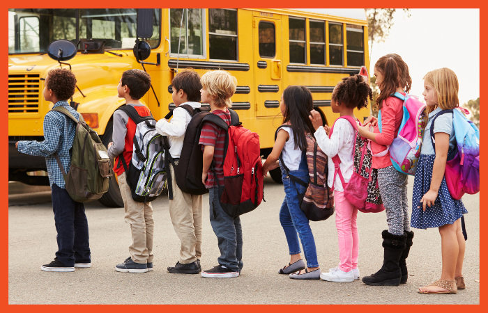 A photo of 8 children standing in front of a schoolbus, all facing left, wearing backpacks.