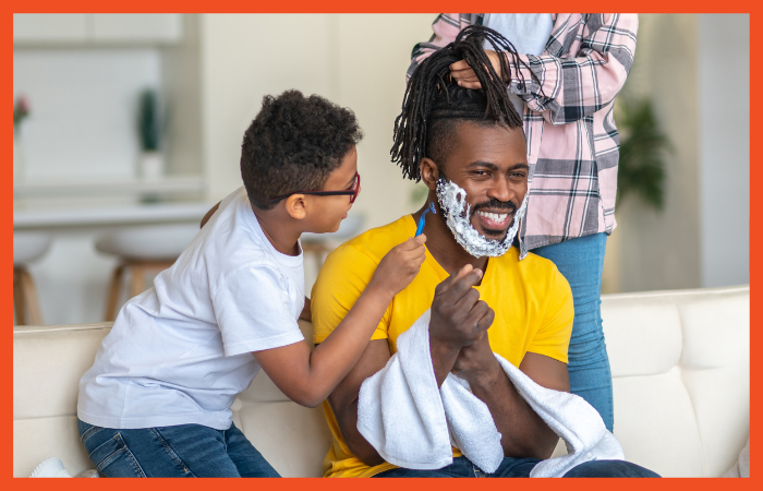 Photo of a Black man sitting on a couch with two children, one playing with his hair and one shaving his face.