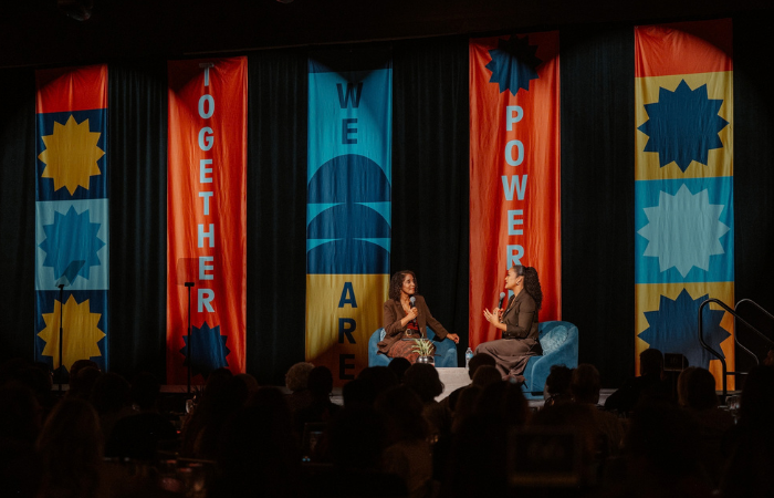 Photo of Angela Poe Russell and Laila Ali together on stage at the 2026 Inspire Luncheon with orange banners behind them that read "Together We Are Powerful"