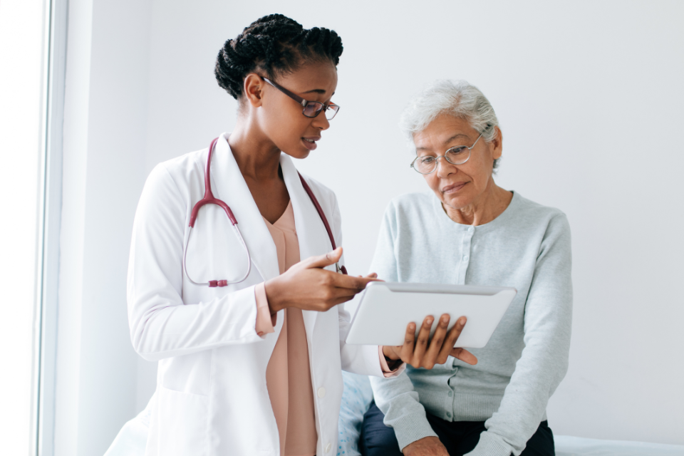 Photo of two Black women in a doctor's office; one elderly, one younger. The younger woman is holding a clipboard to show the older woman, who is sitting down.