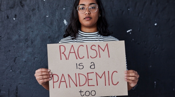 Woman with dark hair, glasses, and brown skin holds a sign that says "racism is a pandemic too"