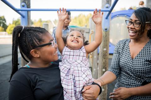 A young girl plays on a jungle gym with the help of her family