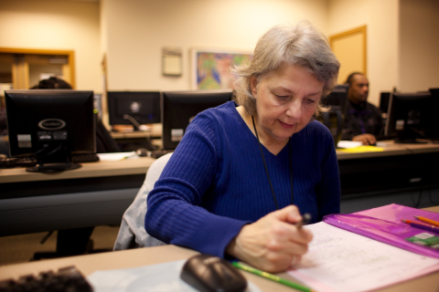 Photo of a woman with grey hair and a blue sweater sitting at a desk writing notes.