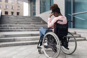 Photo of a young black woman in a wheelchair with one hand resting on her head as she looks at a staircase.